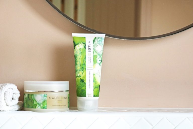 Malée Natural Science 220g Verdure Conditioning Body Scrub 200ml Verdure Conditioning Body Cream placed close to a white rolled towel in front of a half clear mirror with a peach background