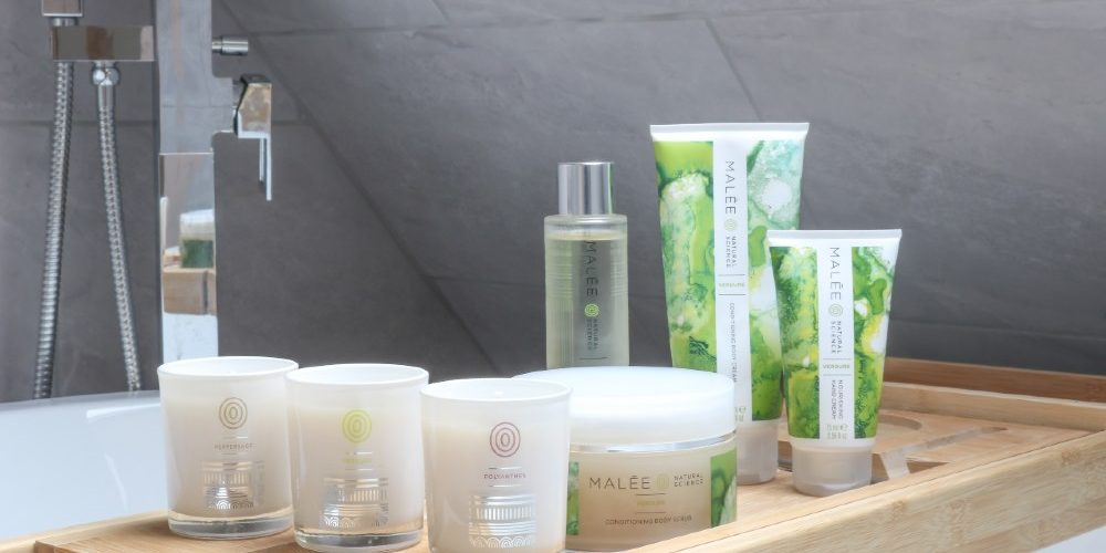 Self-care routine lockdown Malée Natural Science Set on the wooden counter in a bathroom 200ml Verdure Conditioning Body Cream 75ml Verdure Nourishing Hand cream 100ml Verdure Moisturising Oil 250ml Verdure Conditioning Scrub 220ml Malée votives collection