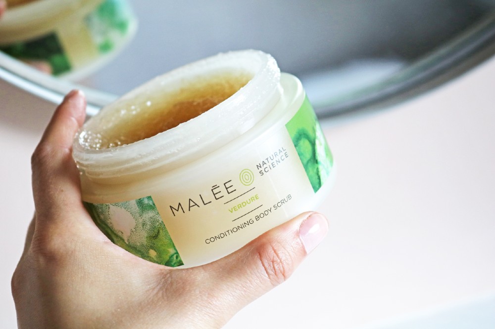 Malée Natural Science a hand holding an opened 220g Verdure Conditioning Body Scrub in front of a mirror 