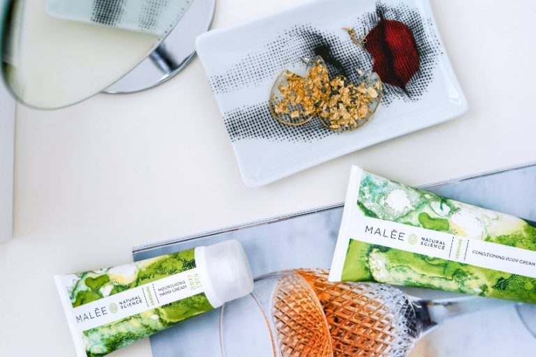 Lips with Lipstick Malee Natural Science On a white table, a glass of wine, 200ml Verdure Nourishing Body Cream, 75ml Verdure Hand Cream, a printed lady nose with a perfect plumped lips with red lipstick napkin, with a pair of golden earrings on the napkin.