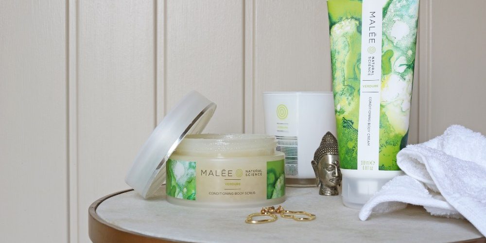 Wellness-Malée-Natural-Science Wellness Malée Natural Science On a side table an opened jar of 250ml Verdure Conditioning Body Scrub 220ml Verdure 100% Soybean Candle 200ml Verdure Conditioning Body Cream a pair of gold hearing and bronze buddha statue with a white towel