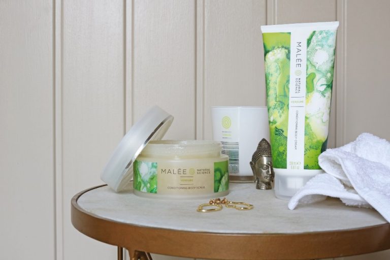 Wellness Malée Natural Science On a side table an opened jar of 250ml Verdure Conditioning Body Scrub 220ml Verdure 100% Soybean Candle 200ml Verdure Conditioning Body Cream a pair of gold hearing and bronze buddha statue with a white towel