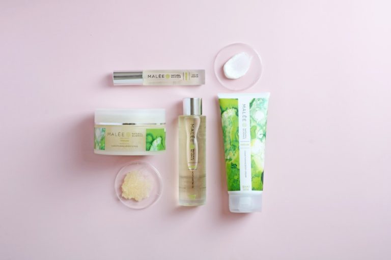 Malée Natural Science two clear lids one pasted with white body cream and the other one with light brown scrub crystals 200ml Verdure Conditioning Body Cream 220g Verdure Conditioning Body Scrub 100ml Verdure Moisturising Oil Verdure Eau De Parfum all together placed on a pink background