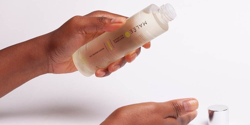 Afro-Hair-Malee-Natural-Science Afro Hair- Malee Natural Science, On a pale pink background, female hands are holding a 100ml Verdure Moisturising oil, with it complete shiny cap next to it.
