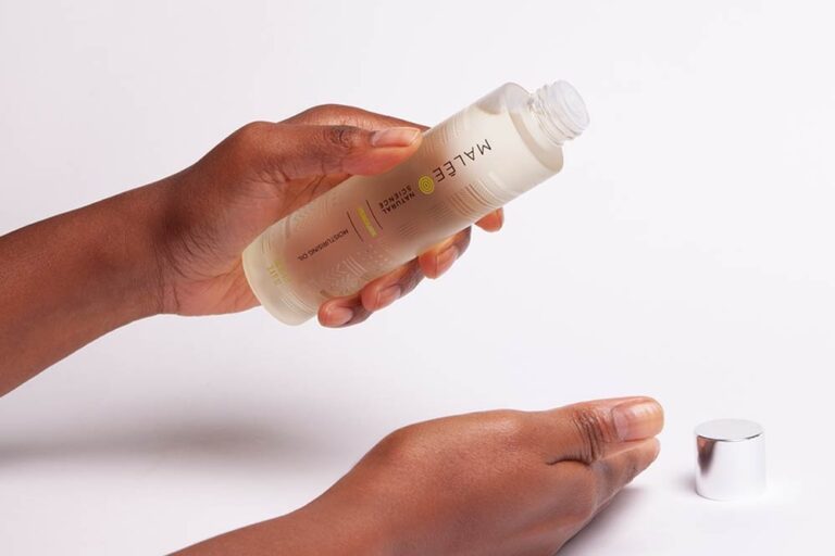 Afro Hair- Malee Natural Science, On a pale pink background, female hands are holding a 100ml Verdure Moisturising oil, with it complete shiny cap next to it.