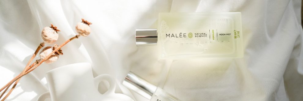 Luxury organic skincare brands Malee Natural Science the 100ml Verdure Room Mist, along with the 12ml Verdure Eau de Parfum are lying side by side on white linen with a white ceramic flower pot and some dry flowers
