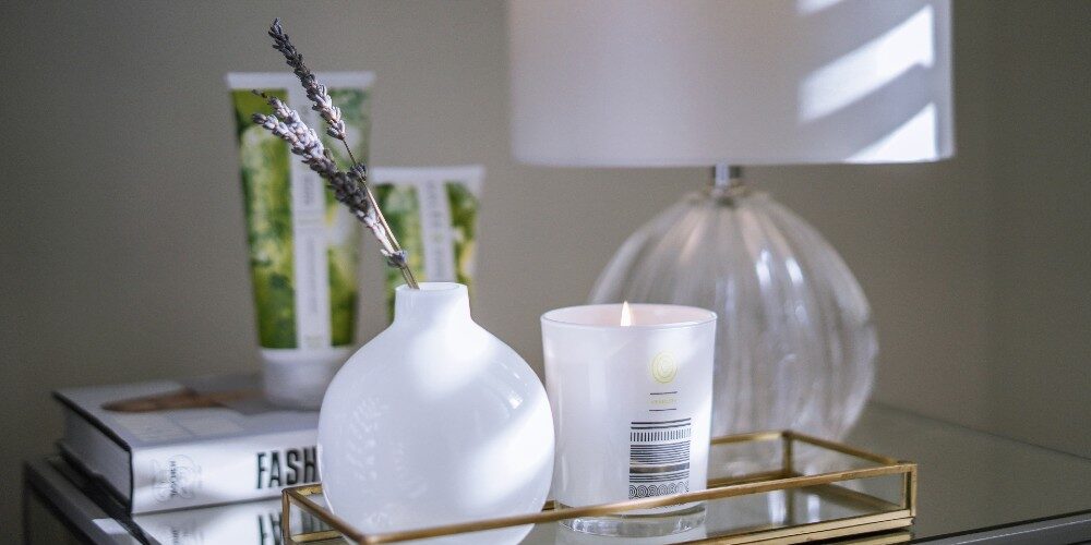 8-Beauty-Routines-Malee-Natural-Science 8 Beauty Routines- Malee Natural Science, on a night table, 200ml Verdure Conditioning Body Cream and 75ml Nourishing Hand Cream are set on a book, next a night light and a golden tray with a lighted 220ml verdure Soy Candle and a white ceramic flowers pot with some lavenders